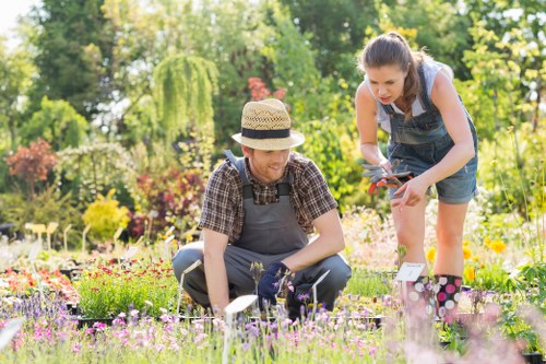 In a well-maintained garden with lush green trees and colorful flowering plants, a man and a young woman are engaged in gardening activity. The man, seated on the grass, wears a straw hat and a checkered shirt under a grey apron, tending to a variety of small flowering plants and shrubs. The woman, standing beside him, is dressed in casual summer clothing with rolled-up jeans and a sleeveless top, leaning forward while holding garden shears. The garden features a neatly edged flower bed with a diverse mix of vibrant pink, purple, and yellow flowers, bordered by grass that appears dense and healthy. In the background, mature trees and bushes provide a natural backdrop, with sunlight filtering through the foliage, indicating a bright, clear day. The environment suggests a peaceful outdoor space suitable for gardening and landscaping services by Landscapers Queens Park, located near Postcode NW6, with a focus on lawn care, plant maintenance, and outdoor aesthetic enhancement.