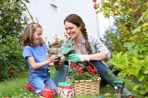 A woman and a young girl are gardening together in a lush, well-maintained backyard. The woman is smiling and wearing green gardening gloves, holding a small potted plant with red flowers, while the girl, dressed in a blue T-shirt and denim shorts, is also engaged in planting or tending to the plant. They are seated on a grassy lawn surrounded by various green plants, shrubbery, and flowering bushes, with some pink flowers visible in the background. Nearby, there are gardening tools including a small green watering can, a red flowerpot, and a wicker basket filled with additional plants, indicating ongoing garden maintenance or planting activity. The outdoor space appears vibrant and healthy, suggesting a well-kept garden that benefits from professional landscaping services such as those offered by Landscapers Queens Park, especially focusing on outdoor gardening and lawn care in the local area. The scene is lit with natural daylight, under clear weather, contributing to the cheerful and productive atmosphere of outdoor gardening in the Queens Park area.