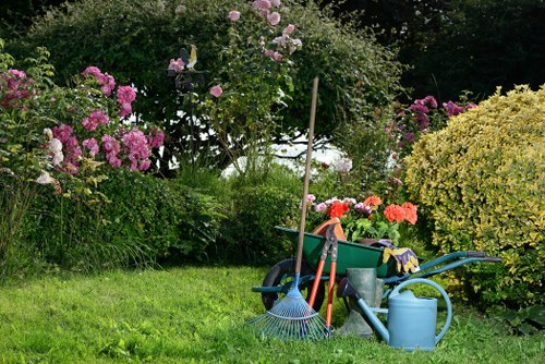 The image depicts a well-maintained garden area with a lush, green lawn in the foreground, bordered by various flowering shrubs and bushes, including pink and purple blossoms on the left and yellow-green foliage on the right. In the background, there are tall trees and dense greenery, creating a natural, tranquil outdoor space typical of a residential garden in Queens Park. The garden tools and supplies, such as a blue watering can, a rake, and a wheelbarrow filled with gardening gloves and small tools, are placed on the grass, indicating recent or ongoing outdoor maintenance. The scene is well-lit with natural daylight, suggesting clear weather conditions that highlight the vivid colors of the plants and the neatness of the lawn. This setting exemplifies a typical landscaped garden, suitable for professional gardening and landscaping services provided by companies like Landscapers Queens Park, with emphasis on lawn care, shrub trimming, and flower bed creation for outdoor enhancement.