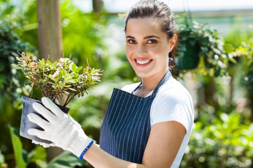 A young woman with dark hair tied back, smiling and wearing a white t-shirt, blue striped apron, and white gardening gloves, holding a potted shrub with small green leaves and pink-tinged new growth in a lush garden environment. The garden features a variety of greenery, including trees and leafy plants in the background, with a wooden post visible to the left. The scene appears to be outdoors on a bright, sunny day, with natural light highlighting the vibrant colors of the plants and the woman's cheerful expression. This setting reflects a well-maintained outdoor space suitable for professional gardening and landscaping services by Landscapers Queens Park, emphasizing healthy plant care and lush garden designs typical of the Queens Park area in London.