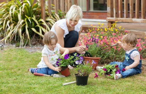 A woman with short blonde hair engaged in gardening alongside two young children in a back garden with a well-maintained lawn. The woman is smiling while helping one child plant purple and pink flowers from a small black pot, with additional flower pots and plants around them. The children, dressed in casual summer clothing, are sitting on the grass and holding garden tools, working on flower planting tasks. In the background, there are lush green bushes, a wooden deck, and a garden fence. The environment appears bright and sunny, with natural lighting highlighting the vibrant colours of the flowers and greenery. The scene exemplifies outdoor gardening activities typical in residential landscaping, aligning with services offered by Landscapers Queens Park in the Queens Park area, NW6 post code, incorporating elements of modern outdoor maintenance and garden enhancement.