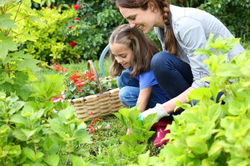 A woman and two young girls are actively engaged in gardening in a lush outdoor space. The garden features a well-maintained, vibrant green lawn in the foreground, surrounded by an assortment of leafy shrubs and plants. In the background, there are additional garden elements such as flowering bushes with red blossoms and a variety of greenery, creating a dense and inviting landscape. The children are focused on a wicker basket, potentially collecting flowers or garden produce, while the woman assists or observes. The scene is set in natural daylight, suggesting good weather, and the environment appears well-kept, reflecting professional gardening or landscaping care by Landscapers Queens Park. The garden's layout emphasizes an organized and lush outdoor area suitable for outdoor activities and plant care, consistent with gardens in the Queens Park region, London, SW10 postcode area.