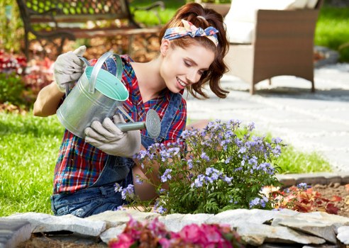 A woman with light skin and brown hair tied back with a colorful headband is kneeling in a landscaped garden, watering a cluster of light purple flowering plants using a small metal watering can. She is dressed casually in a checkered shirt and denim jeans, and is wearing gardening gloves. The garden features a curved stone border surrounding the flower bed, with various plants and flowers in the foreground, including some pink blooms. In the background, there is a wooden outdoor dining set with a table and chairs on a paved patio, surrounded by lush green grass, trees, and shrubs, suggesting a well-maintained residential garden in Queens Park. The scene is illuminated by natural daylight, indicating a clear and sunny day, and the overall environment reflects a tidy and vibrant outdoor space that benefits from professional gardening and landscaping care from companies like Landscapers Queens Park.