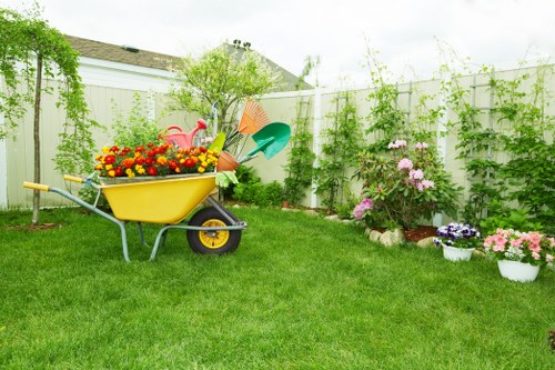 A well-maintained backyard garden with a lush, green lawn occupying the foreground, bordered by vibrant flower beds containing blooming pink and purple flowers in white pots and planting borders. In the background, a tall, white wooden fence encloses the space, with climbing green plants and vines growing along it, contributing to a natural and lush appearance. To the left, a bright yellow wheelbarrow filled with gardening tools such as watering cans, trowels, and colorful plastic equipment is positioned on the grass, indicating recent or ongoing garden maintenance. A small, leafy tree with fresh green foliage stands behind the wheelbarrow, adding vertical interest to the scene. The garden appears to be outdoors on a partly cloudy day, with soft, natural light illuminating the space, ideal for gardening activities. The overall environment suggests a peaceful, cared-for outdoor area suitable for gardening and landscaping services offered by Landscapers Queens Park, set within a typical UK suburban garden setting with a focus on flower and plant care.
