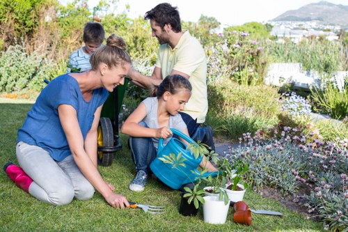 A family engaging in gardening activities in a well-maintained backyard garden with lush green grass, a variety of flowering plants, and shrubs. The foreground features two children and an adult woman kneeling on the lawn, planting or tending to the soil, with gardening tools and plant pots nearby. The children are dressed casually, one holding a small gardening trowel, while the woman assists with the planting. In the background, a man with dark hair and a beard stands smiling, overseeing the activity, with additional garden beds filled with blooming flowers and hedges visible. The garden is bordered by a mix of greenery and vibrant flowers, and the environment appears to be in bright daylight with natural sunlight, suggesting a clear, pleasant day. This scene illustrates outdoor garden maintenance and family-focused gardening, typical of landscaped outdoor spaces managed by local UK landscapers such as Landscapers Queens Park, emphasizing lawn care, planting, and outdoor family activities within a tidy and inviting garden setting in the Queens Park area.