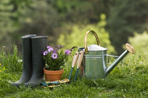 An outdoor garden scene featuring a pair of black rubber gardening boots standing on lush green grass, positioned next to a terracotta pot filled with purple and white flowers. Nearby, a collection of garden tools with wooden handles, such as pruning shears and a hand trowel, are arranged on the grass. A metal watering can with a long spout and a curved handle is also present, sitting amidst the tools and plant container. The background reveals a softly blurred natural landscape with trees and foliage, suggesting a well-maintained, landscaped garden area typical of outdoor gardening spaces in Queens Park. The scene is illuminated by natural daylight, indicating mild weather conditions suitable for gardening activities, and reflects an environment indicative of lawn and garden care offerings provided by Landscapers Queens Park, aligned with general gardening and landscaping services in the local area.