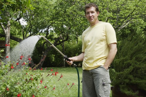 A man wearing a pale yellow t-shirt and grey cargo shorts is standing in a well-maintained garden in Queens Park, watering a variety of flowering plants and shrubs with a garden hose. The garden features a lush, green lawn with a smooth, even surface, bordered by vibrant flower beds filled with red blooms. In the background, dense trees and neatly trimmed hedges create a natural enclosure, while a paved patio area and a large, leafy shrub add structure and depth to the outdoor space. The scene is set on a bright, partly cloudy day, with natural sunlight illuminating the greenery and highlighting the fresh, natural tones of the garden. This setting exemplifies professional gardening and landscaping maintenance, emphasizing healthy plant growth and well-organized outdoor environments, similar to services provided by companies like Landscapers Queens Park.