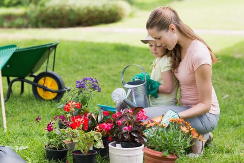 A woman and a young girl are engaged in gardening activities in a well-maintained outdoor garden, which features a green lawn with neatly trimmed grass, an assortment of potted flowering plants with vibrant red, purple, and orange blooms, and several garden tools and watering cans placed nearby. In the background, a garden wheelbarrow with yellow wheels is positioned on the grass, and a row of shrubs and mature trees provide natural screening and a lush environment. The scene is set in daylight with soft natural lighting, indicating pleasant weather suitable for outdoor gardening. The garden layout includes a mix of flower beds and open grassy areas, reflecting a landscaped yard maintained by local gardening specialists such as Landscapers Queens Park. The woman is wearing a casual pink T-shirt and jeans, and the girl has on a yellow top and green gloves, showing an engaging and family-friendly gardening activity that supports outdoor landscaping and lawn care services. The scene captures a peaceful and productive moment of gardening, highlighting the natural textures of soil, plants, and grass, and underlines the importance of garden maintenance in a residential outdoor setting near Queens Park.