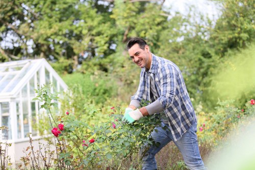 A man in a blue checked shirt and jeans is smiling while tending to flowering shrubs in a well-maintained garden, which features a lush green lawn, neatly edged flower beds with pink and red blooms, and a white greenhouse structure in the background. The garden is surrounded by mature trees with dense foliage, creating a peaceful outdoor environment under clear weather conditions. The scene highlights garden maintenance activities typical of landscaping services in the Queens Park area, with natural tones of green, pink, and brown, and a focus on outdoor plant care and tidy landscaping features.