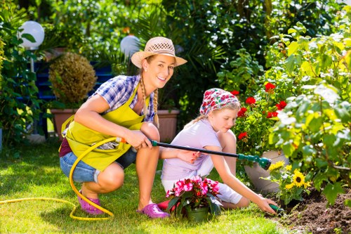 A woman and a young girl working together in a lush garden, with the woman wearing a straw hat, yellow apron, and casual clothing, while the girl is dressed in a floral hat and white outfit. They are tending to a flower bed surrounded by blooming red, yellow, and pink flowers, with the woman holding a garden hose and the girl planting or inspecting the soil. The garden features a neatly maintained lawn, dense green shrubs, and a variety of flowering plants, with decorative lanterns placed along a fence or backing hedge. The scene is brightly lit by natural sunlight, suggesting a warm, sunny day, and the setting appears to be a well-kept, residential outdoor space in Queens Park, London, suitable for outdoor gardening activities and family gardening projects.